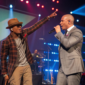 Ne-Yo and Pitbull performing on stage. Ne-Yo, in brown hat and plaid shirt, gestures dramatically. Pitbull, in gray suit, sings into microphone. Stage lit with vibrant lights, surrounded by equipment and illuminated backdrop.