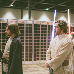 Actors standing at a sci-fi convention. Backdrop features Star Wars banners and iconic lightsabers.