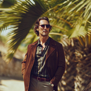 Matthew McConaughey, in a casual yet stylish outfit, posing on a sunny beach. Palm trees sway in the background.