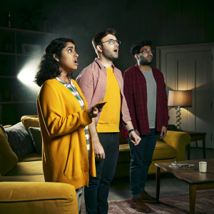 Geraldine Viswanathan, Daniel Radcliffe, and Karan Soni standing in a tense moment from a popular sitcom. The scene is set in a cozy living room with soft lighting and comfortable furniture.