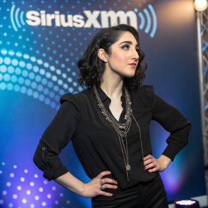 Emily Estefan in an elegant black blouse and necklace. Confident pose against a dynamic backdrop featuring the SiriuxXM logo. Vibrant colors, studio lighting.