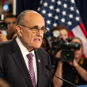 Rudy Giuliani, middle-aged man, stern look, gray hair, glasses, dark suit. Press conference backdrop