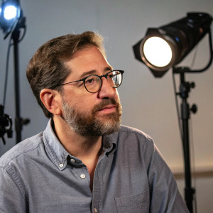 Glenn Greenwald, with a beard and glasses, dressed in a casual shirt. Appearing on a podcast, surrounded by studio lights.