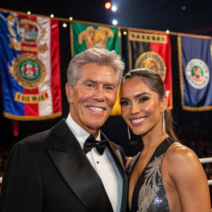 Michael Buffer and Christine Prado at a boxing gala. Both are beaming with joy, set against a backdrop of boxing championship banners.