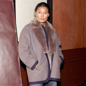 Song Haena, Asian model, standing against a stark black backdrop. Wearing a oversized gray coat with fur collar. Solemn expression. Studio photo