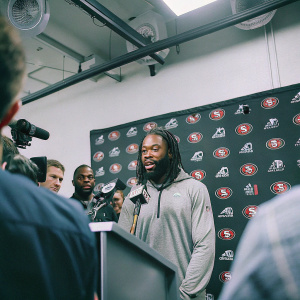 Richard Sherman, bearded NFL player, speaking at a press conference. The backdrop is filled with the San Francisco 49ers and NFL logos. Indoor setting with multiple reporters and cameras.