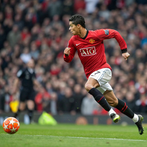 Cristiano Ronaldo, wearing the iconic Manchester United jersey adorned with AIG logo, sprinting during a crucial match. The crowd is a blur in the background. A glowing orange ball is inches away from his feet.
