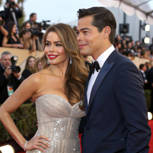 Sofia Vergara and Manolo Gonzalez-Ripoll Vergara at an award show. Sofia in a shimmering silver gown. Manolo in a navy suit. Crowded room with flashing cameras.