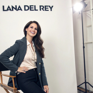 Lana Del Rey on the set of a promotional shoot, smiling against a stark white backdrop with sponsors' logos.