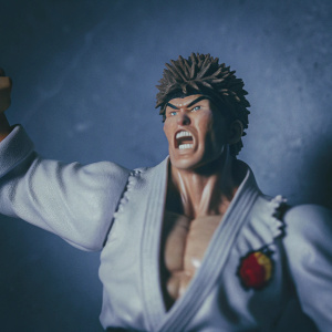 Miniature figurine of Ryu from Street Fighter. Intense expression, fiery emblem on chest. Dark background, dramatic pose with arms outstretched