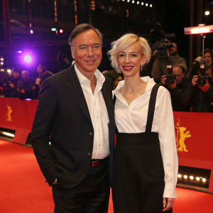 Nico Hofmann and Katja Eichinger at the Berlinale gala. Wearing formal attire. Dark blazer, white shirt for Nico. White shirt and black dress for Katja. Standing on red carpet. Bright lights and paparazzi in background.
