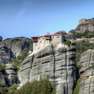 Meteora monastery perched on a massive rock formation in Greece. The ancient Greek architecture with its stone facade and red-tiled roofs stands out against the backdrop of rugged, layered rocks covered in lush greenery. The blue sky and bright sun illuminate the scene, creating a vibrant and realistic image highlighting the rock formations and vegetation.