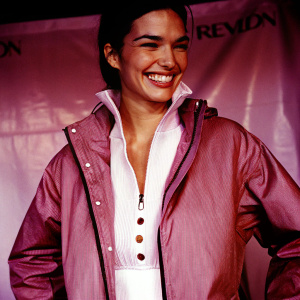 Karen Duffy, smiling broadly, in a red jacket with Revlon logo. White blouse underneath. Dark fabric backdrop with text. High-resolution photo.