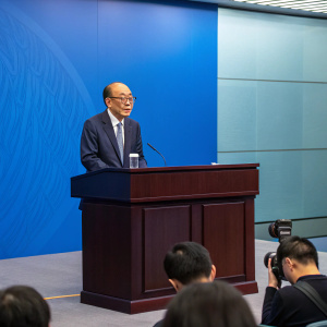 Li Ka-shing, business tycoon, speaking at a press conference. Modern office setting, dark wooden table, blue backdrop