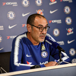 Maurizio Sarri, Chelsea FC manager, during a press conference. Seated behind a desk, he wears a blue and white club jersey. Focused expression as he addresses the media.