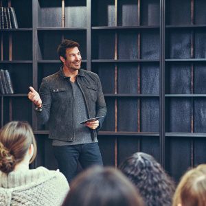 Dylan McDermott at a book signing event. Wearing a casual outfit. Dark wooden bookshelf backdrop.