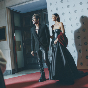Jon Kortajarena and Lily Aldridge at the Met Gala. Jon in black suit with leather pants and boots. Lily in strapless gown. Confident poses. Logos and graphics on white backdrop.