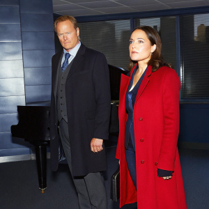 Ulrich Thomsen and Megan Boone in a tense scene from 'The Blacklist'. Dark, moody atmosphere with dim lighting and a grand piano in the background.