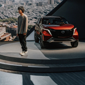 Concept car reveal event featuring Nissan Gripz. The vehicle, a vibrant red and black hybrid, stands on a circular pedestal. A youthful Asian male model, dressed in casual attire, poses beside the car. The backdrop showcases a digital representation of a bustling cityscape.