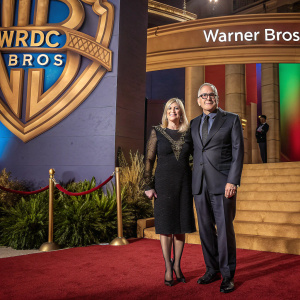 Penny Coulter and Jim Coulter at a high-profile gala. Penny in a black dress with detailed embroidery, Jim in a tailored dark suit. Behind them, a vibrant backdrop with logos of NRDC, BROs, and WARNER BROS.
