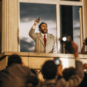 Fidel Castro addressing a crowd from the balcony. Dressed in gray suit, white shirt, red tie. The evening sky provides a dramatic backdrop.