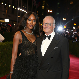 Naomi Campbell and Manolo Blahnik walking the Met Gala red carpet. Formal attire. The backdrop is illuminated by bright city lights.