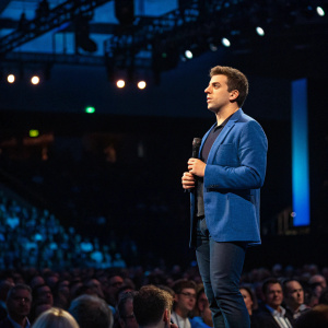 Brian Chesky, standing on stage during a tech conference, wearing a blue blazer. Crowded audience in the background