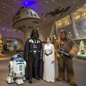 Darth Vader, Princess Leia, Han Solo, R2-D2, and Chewbacca pose for a publicity shot. The setting is a vibrant, space-themed exhibition hall with starship models.