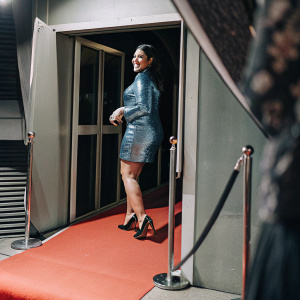 Janina Gavankar, south asian actress, confidently posing in a shimmering blue dress. Black heels. Promotional backdrop for a blockbuster movie, red carpet event.