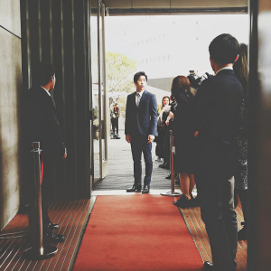 Song Joong-ki, wearing a navy suit, stands on the blue carpet. Event attendees and photographers in the background