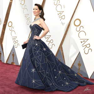 Lucy Liu on the Oscars red carpet. Wearing an elegant dark blue gown with geometric patterns. Confident pose. White background with event logos