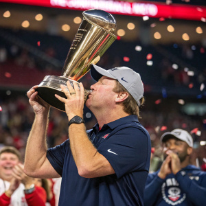 Coach Kirby Smart celebrating victory with a kiss on the gleaming trophy. Wearing a navy polo and baseball cap. Stadium crowd cheering in the background