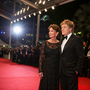 Robert Redford and Sibylle Szaggars at Cannes Film Festival. Dressed in black, standing on a red carpet. Bright lights and crowds in background.