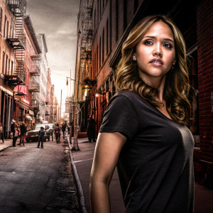 Jessica Alba, dressed in a casual black t-shirt, standing against a busy city street backdrop. She has a light makeup look and long wavy brown hair.