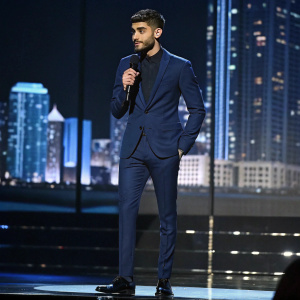 Zayn Malik at a gala event. Wearing a navy blue suit, he stands confidently on a stage with a cityscape backdrop.