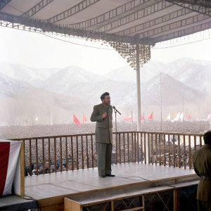 Fidel Castro in a formal suit, addressing a large crowd at a political rally. Behind him, a stage with banners and a backdrop of mountains.