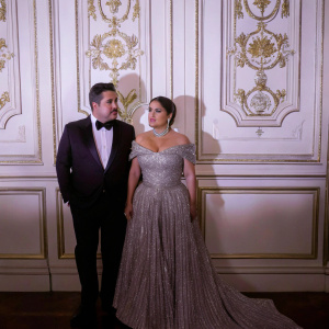 Salma Hayek and Narciso Rodriguez at the Met Gala. Salma in a shimmering off-the-shoulder gown, Narciso in a tuxedo. White backdrop with intricate gold logos.