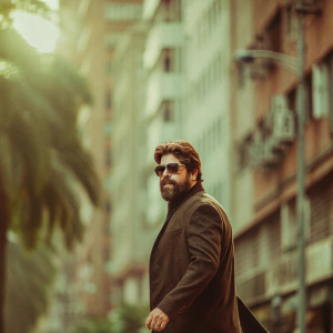 Benicio del Toro walking down a bustling city street. Full beard and styled hair. Dark tailored jacket. Sunlight filtering through tall buildings creating soft shadows. Background blurred, focus on del Toro.