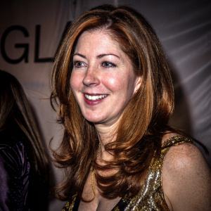 Dana Delany, middle-aged actress with a warm smile, in a gold detailed dress. Candid pose, upper body focus. White fabric backdrop with GL letters.