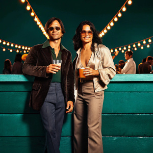 Gael Garcia Bernal and Michelle Rodriguez at the Cannes Film Festival. Posing with drinks in hand, vibrant festival lights and bustling crowd in the background.