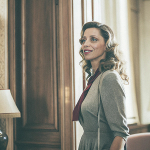 Angela Sarafyan, Caucasian woman, with warm eyeshadow, soft makeup, loose voluminous curls, wearing gray dress and maroon accessory. She stands in a luxurious office space with rich wood paneling.
