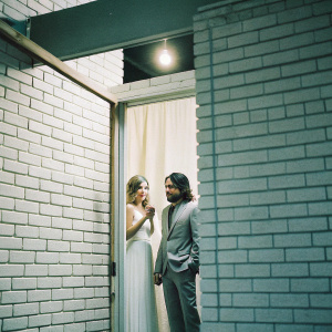 Zoey Deutch and Avan Jogia at a glamorous gala. Zoey in a white gown, Avan in a grey suit. Intimate setting with warm lighting, brick wall backdrop.