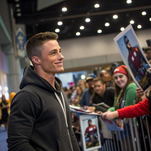 Colton Haynes, dressed in a casual hoodie, at a comic-con event. Behind him, fans holding posters of 'San Andreas'