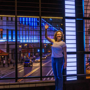 Scarlett Johansson, smiling and waving at fans. Dressed in a casual white t-shirt and blue jeans. Background is a bright sunny day on a bustling city street.