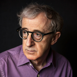 Close-up of Woody Allen, wearing a purple shirt and glasses. Serious expression. Dark background