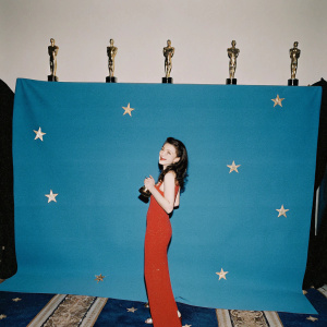 Cher Lloyd, wearing a vibrant red dress, at a glamorous awards show. Golden statues and stars in the background.