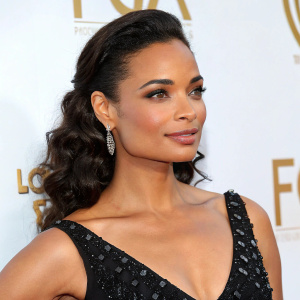 Rochelle Aytes at a gala event. Elegant black dress with sequins. White backdrop with gold accents. Close-up shot focusing on her face and upper body