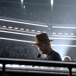 Justin Timberlake performing live at the Grammy Awards. Wearing a brown fedora and dark t-shirt. Flashing lights and crowd in background.
