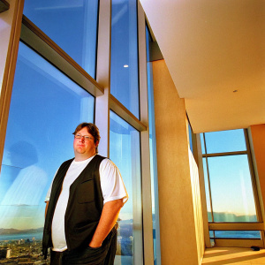 Gabe Newell, standing in Valve Corporation's office, wearing a black vest over a white t-shirt. Serious expression. Modern office setting with glass windows overlooking a cityscape.
