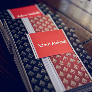Stacks of business cards for Adorn Mehndi. Bold red and black design. Sans-serif font. Professional, organized look. Close-up shot.